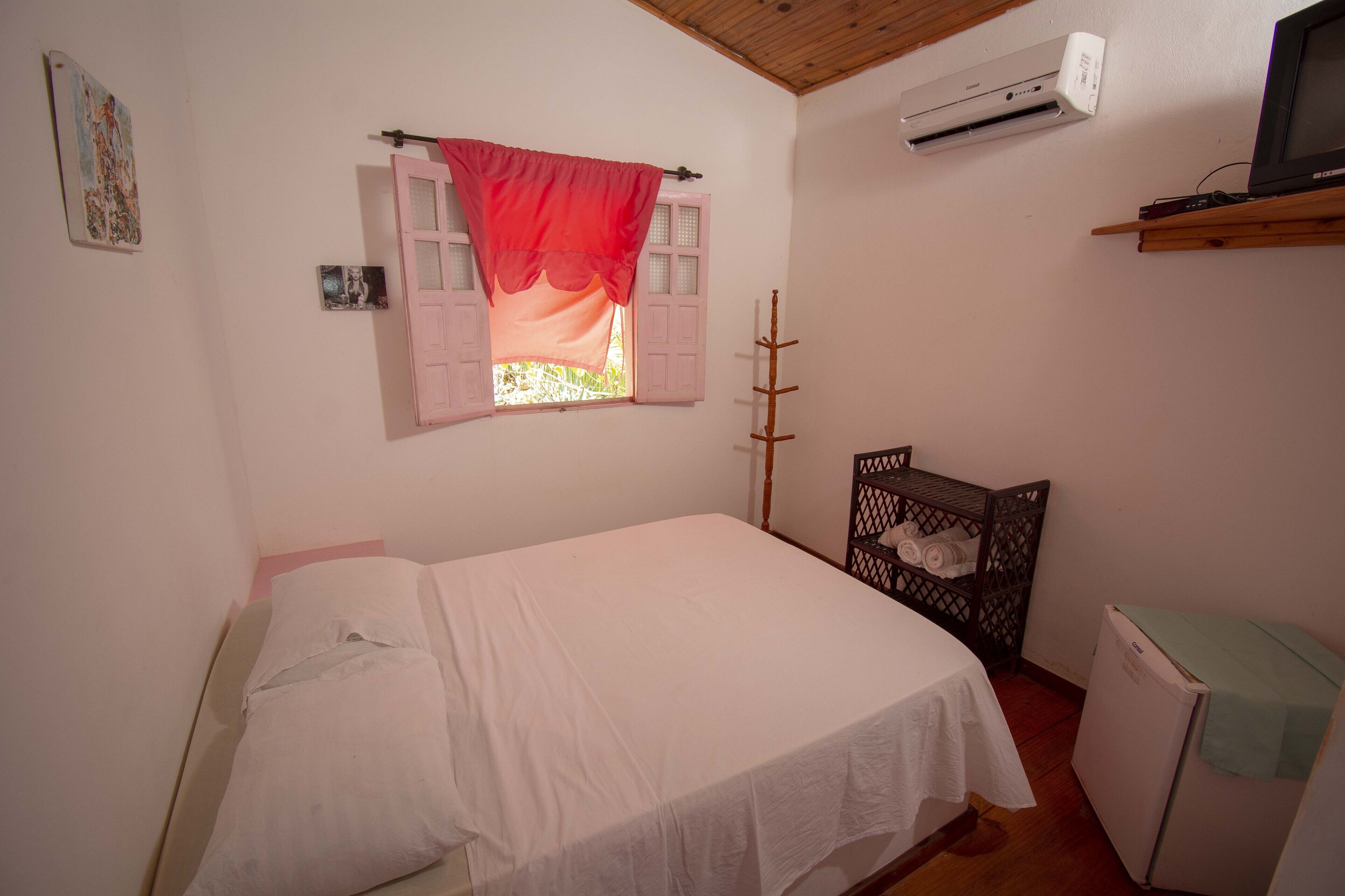 Ein Schlafzimmer mit einem Einzelbett, einem Fenster mit roten Vorhängen, einer Klimaanlage, einem Fernseher auf einem Holzregal und einem kleinen Kühlschrank.