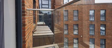 A modern balcony with a wooden floor, glass railing, and a view of adjacent brick buildings.