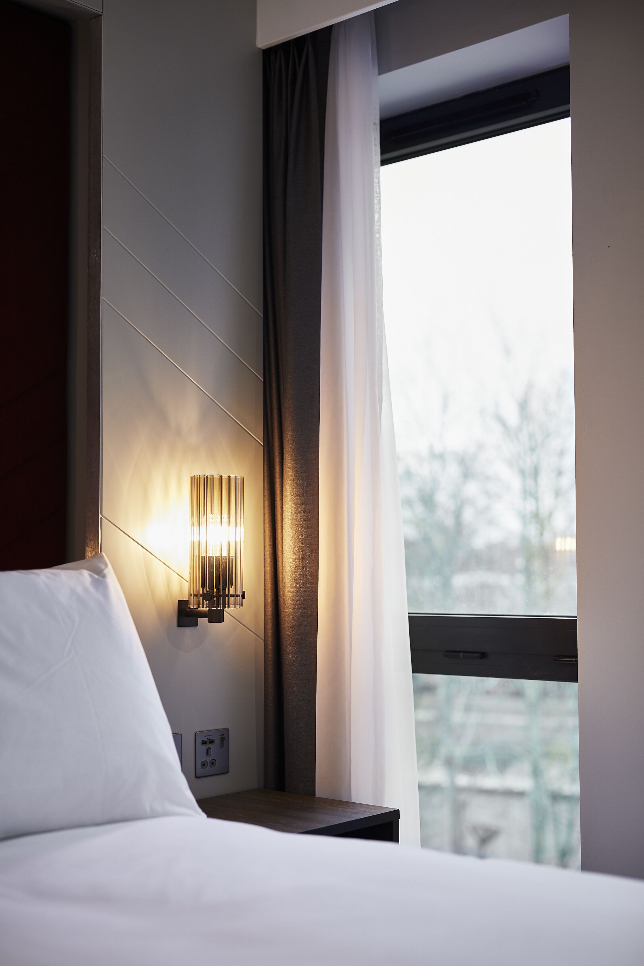 Een hotelkamer met een bed, een wandlamp, een raam met gordijnen en uitzicht op bomen.