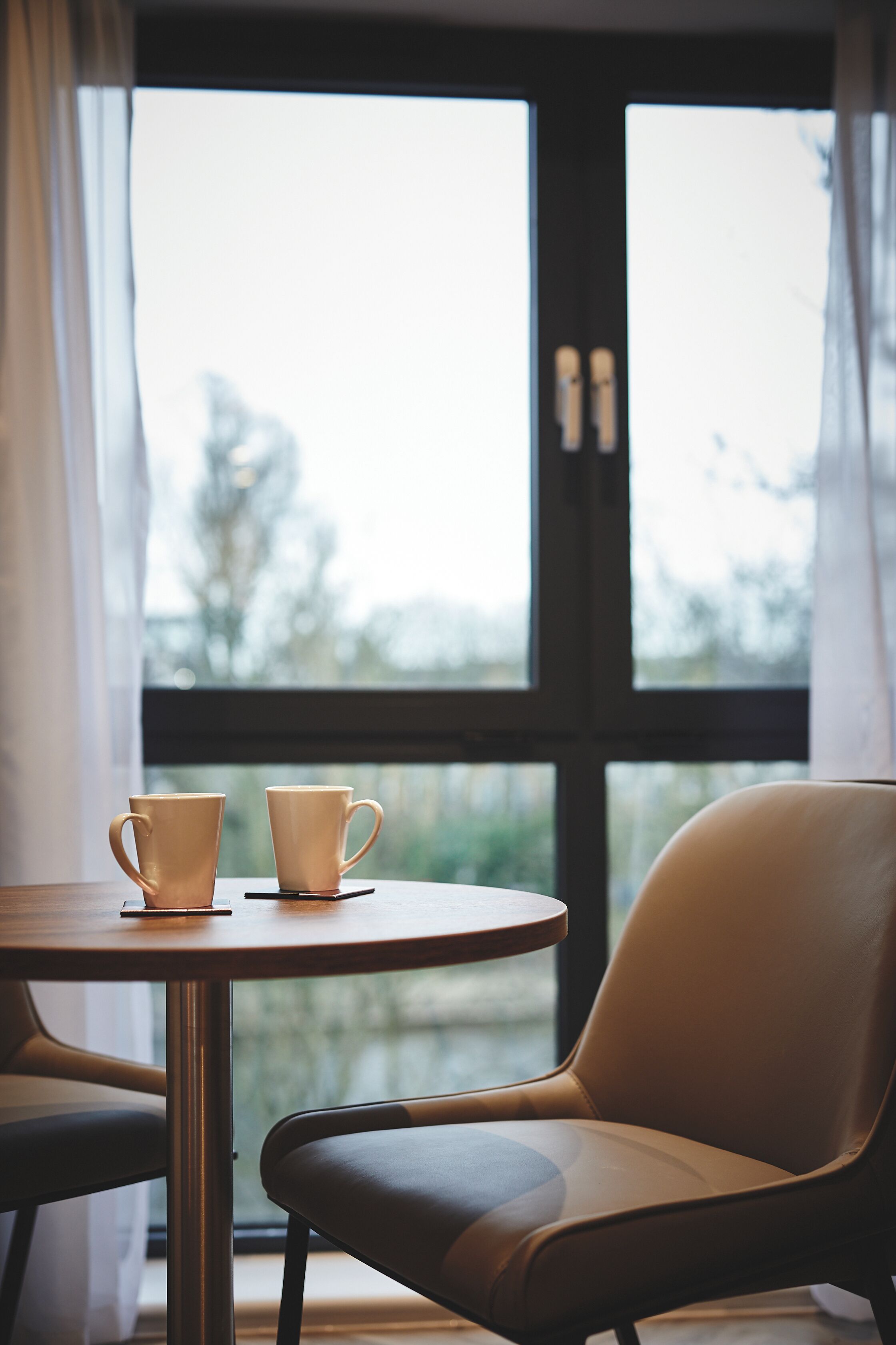 Une table avec deux tasses et une chaise devant une fenêtre ornée de rideaux.