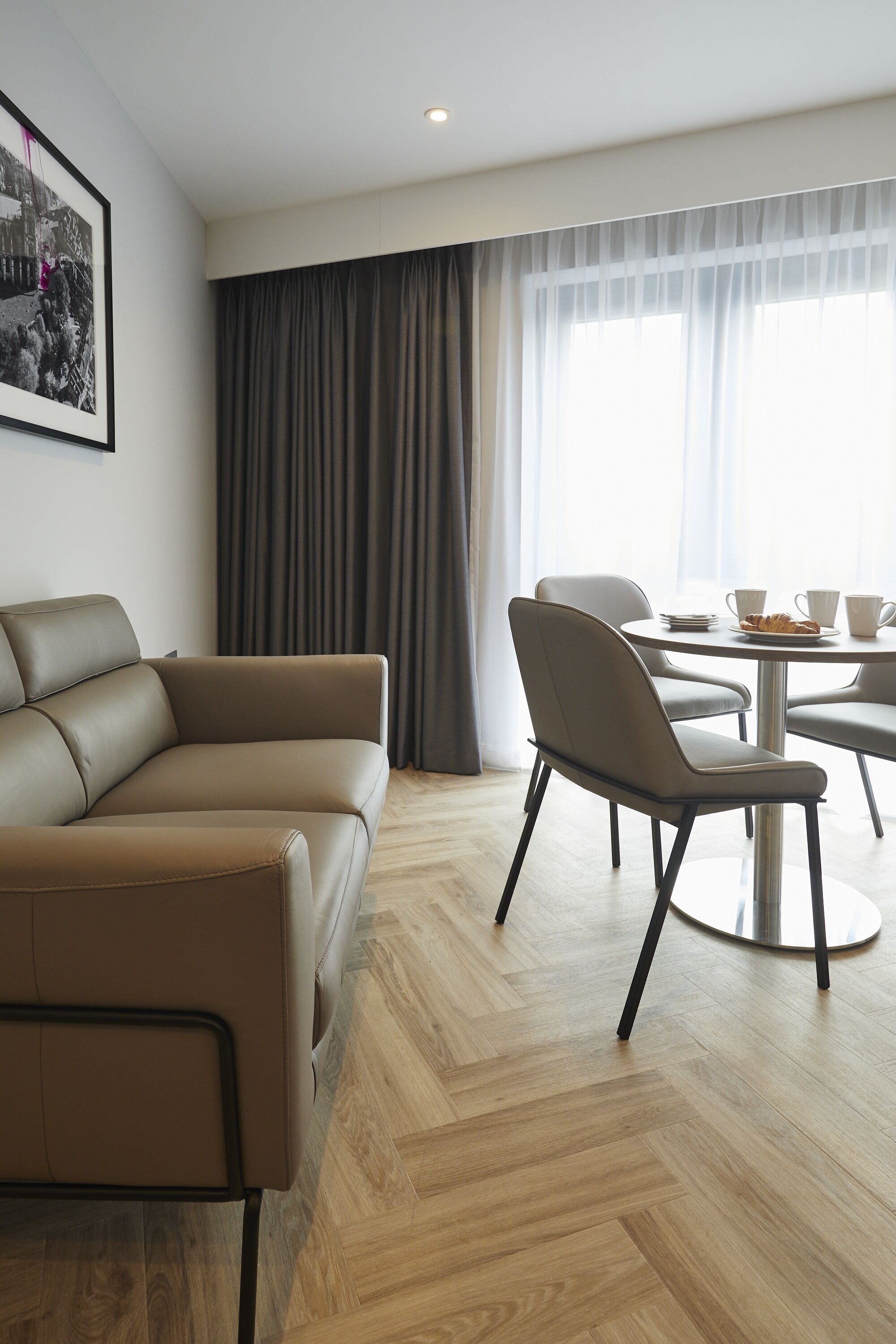 A modern living room with a brown leather sofa, a round dining table with chairs, a large window with sheer curtains, and a framed picture on the wall.