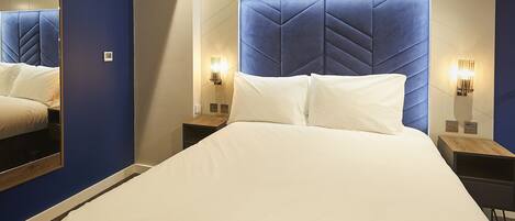 A modern hotel room with a large bed, a headboard with geometric patterns, a nightstand with a lamp, and a wall-mounted mirror.