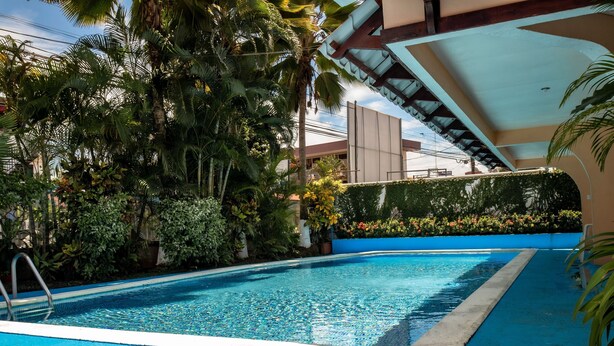 A swimming pool with a blue edge, surrounded by lush greenery and a covered area with a sloped roof. - Hotel y Cabinas El Coral (Jacó)