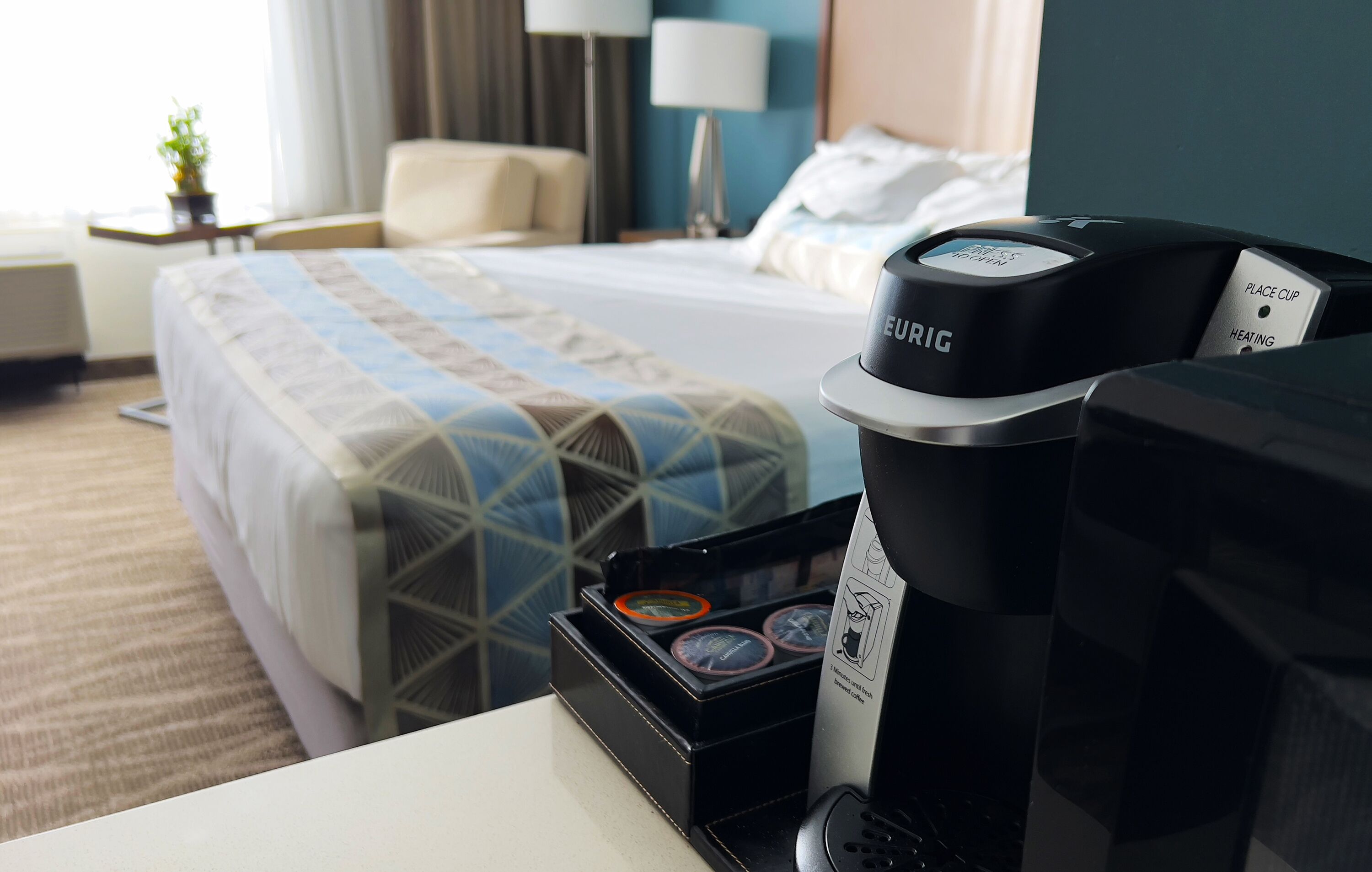 A hotel room with a bed, a desk with a coffee maker, and a nightstand with a lamp.