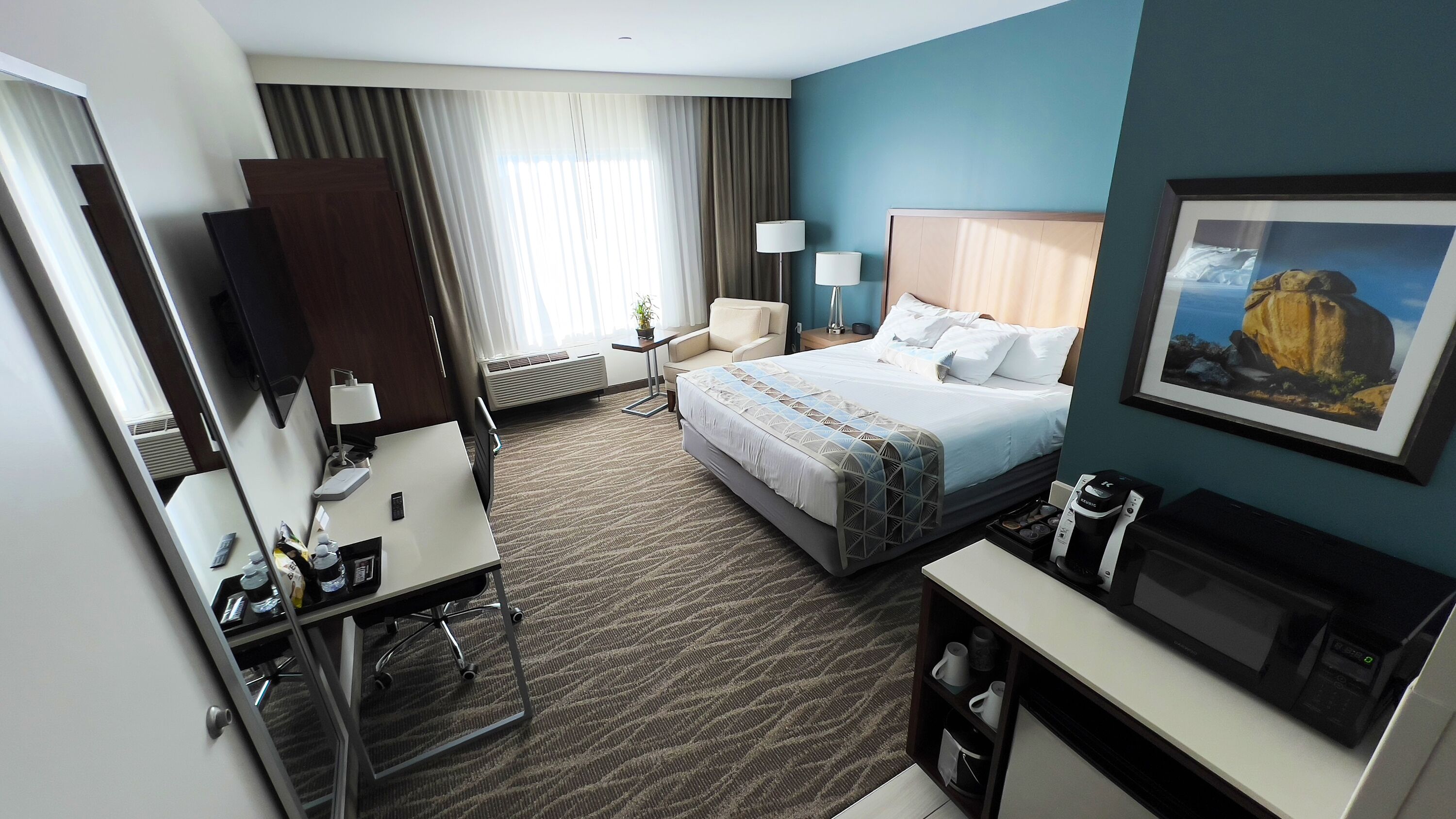 A hotel room with a bed, desk, chair, TV, and a painting on the wall.