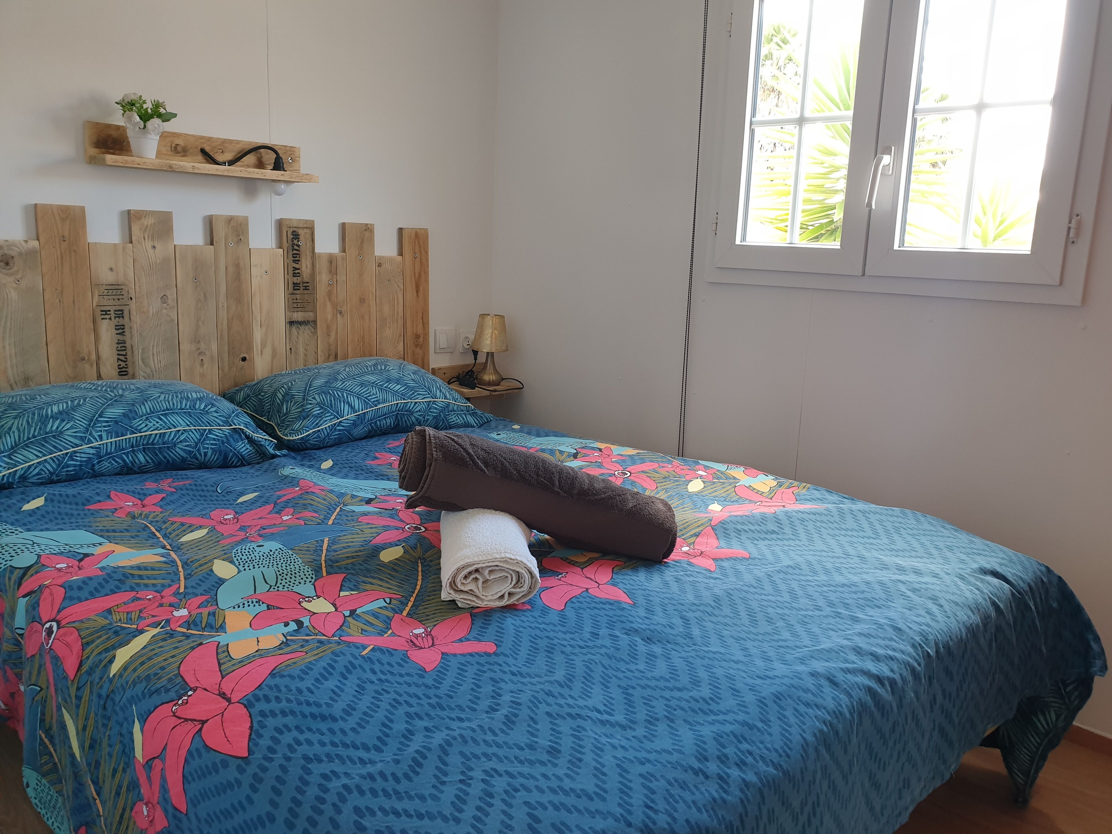 A bedroom with a wooden headboard, a floral bedspread, and a window with a view of greenery.