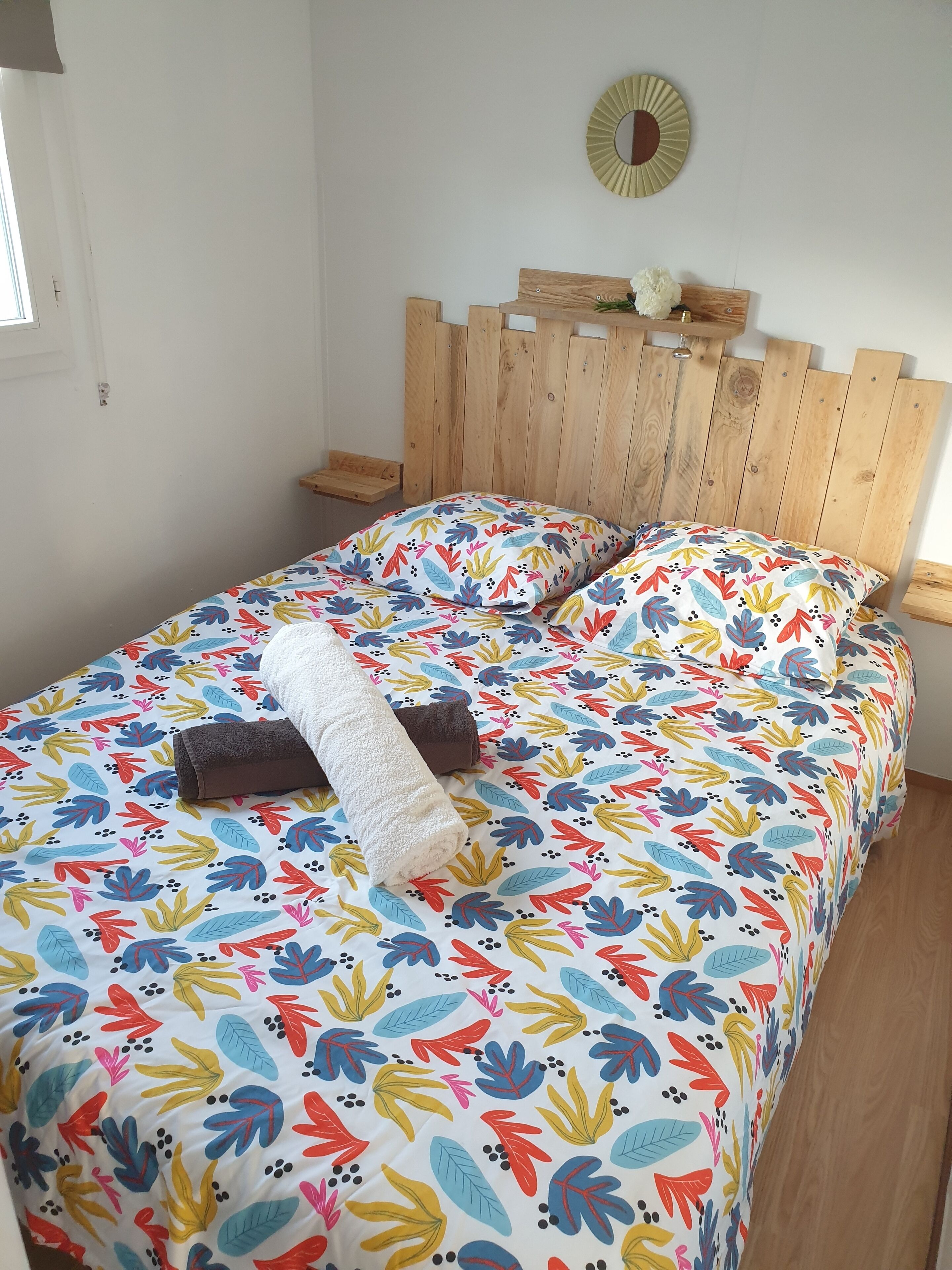 Una cama con una colcha estampada de flores, un cabecero de madera y una pequeña estantería con un objeto decorativo.