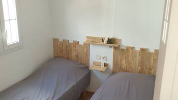 A small, clean room with two single beds, a wooden headboard, a small shelf with a speaker, and a window.
