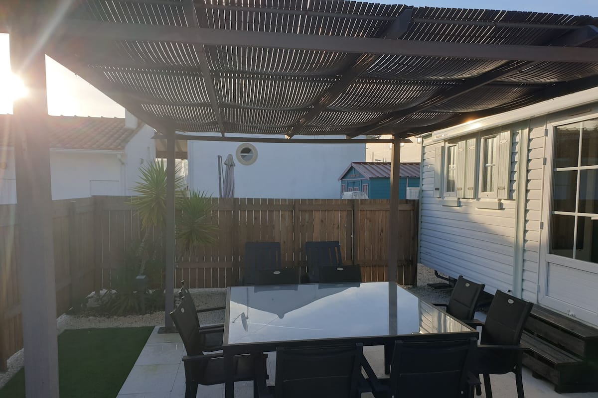 Une terrasse dotée d’une table à plateau en verre et de chaises noires, entourée d’une clôture en bois et avec une maison en arrière-plan.