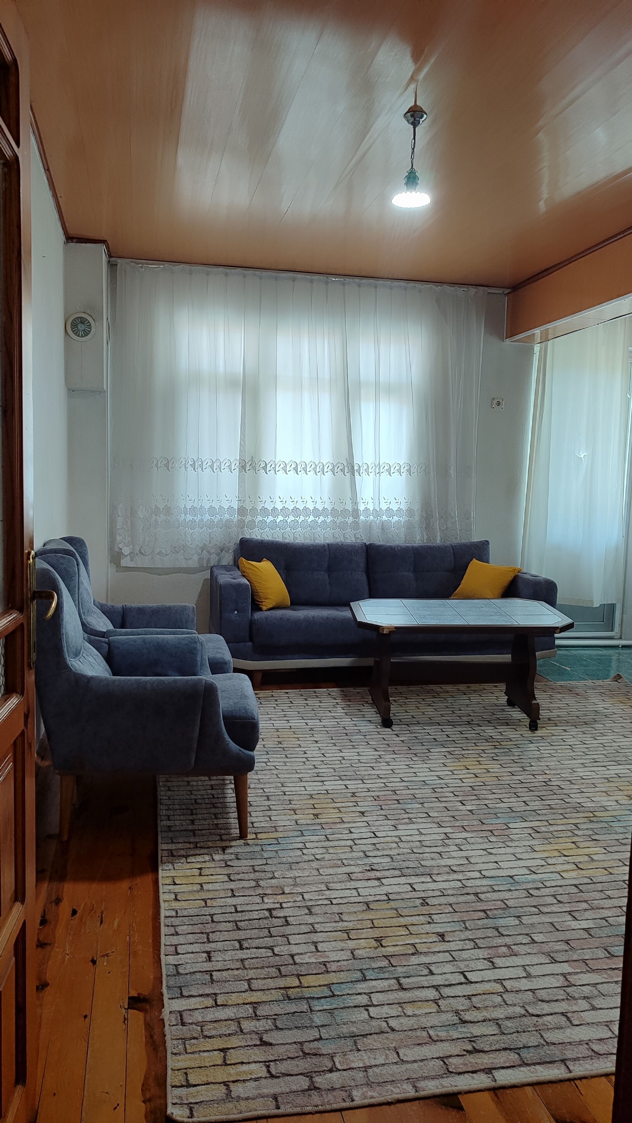 A room with a sofa, two armchairs, a coffee table, a patterned rug, wooden flooring, and a ceiling fan.