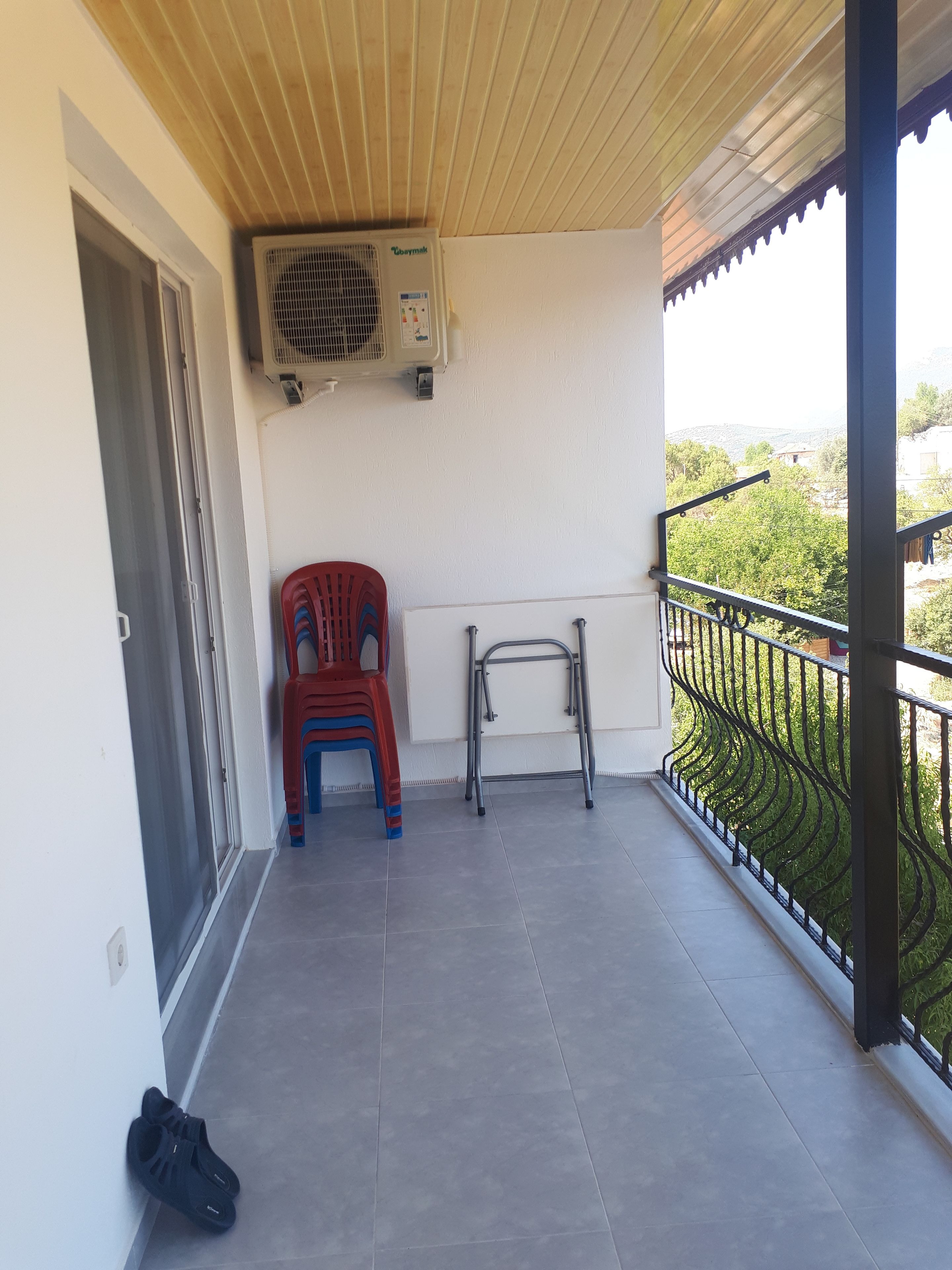 A balcony with a tiled floor, a red and blue plastic chair, a metal folding chair, and a pair of black shoes.