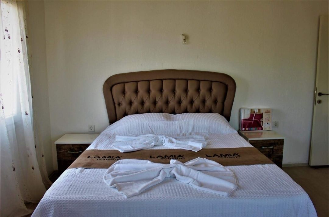 A neatly made bed with a tufted headboard, a white bedspread, and a brown towel with "LAMIA" branding. There are two bedside tables with a red object on one.