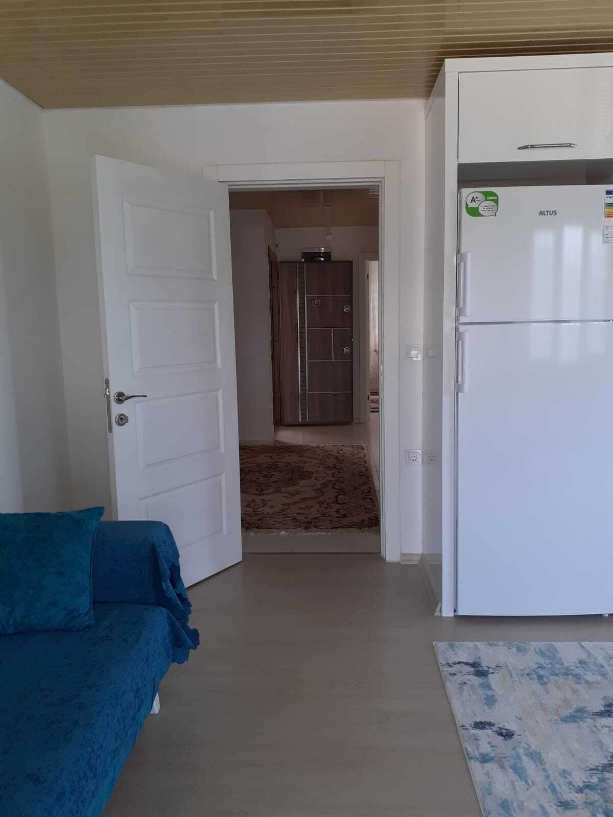 A room with a white door, a blue sofa, a white refrigerator, and a shower area in the background.