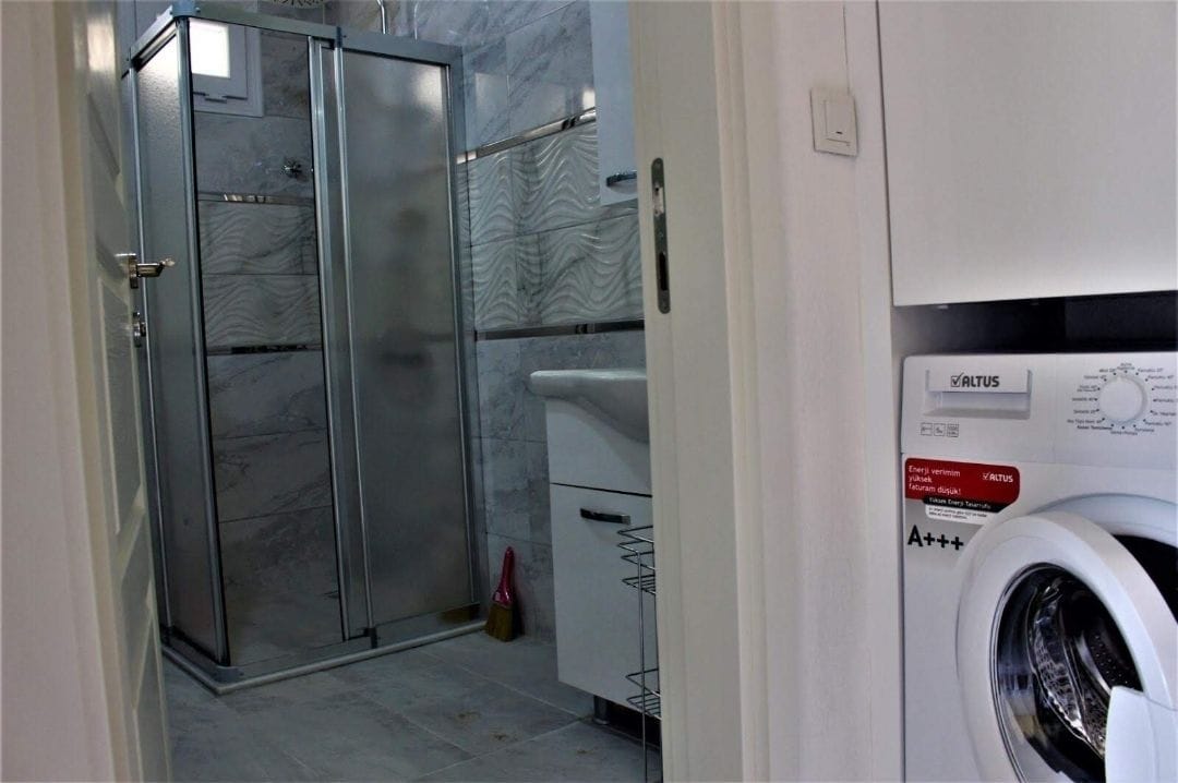 A compact bathroom with a glass shower enclosure, a built-in washing machine, and a tiled floor.