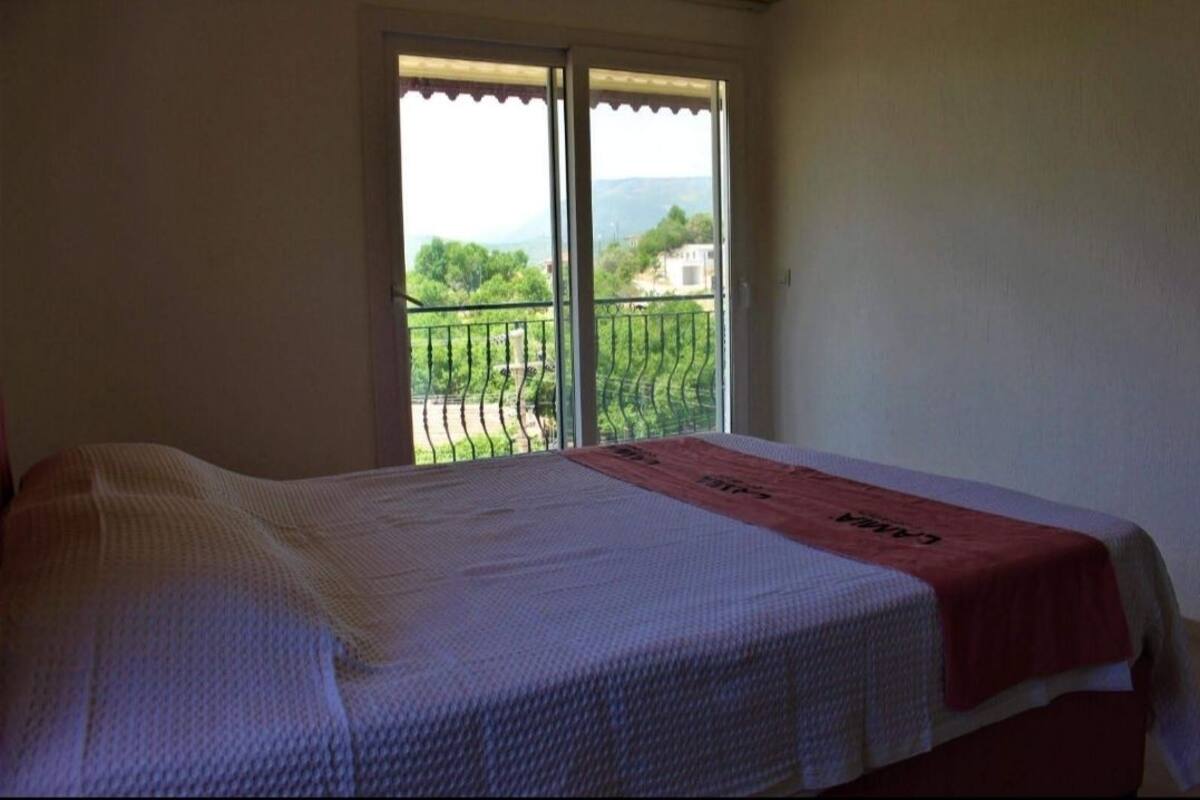 A bedroom with a bed, a red towel, and a balcony with a view of greenery and buildings.