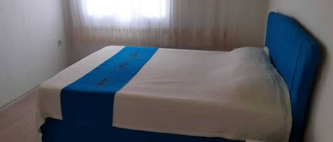 Una habitación pequeña con cama individual, cabecera azul y colcha blanca.