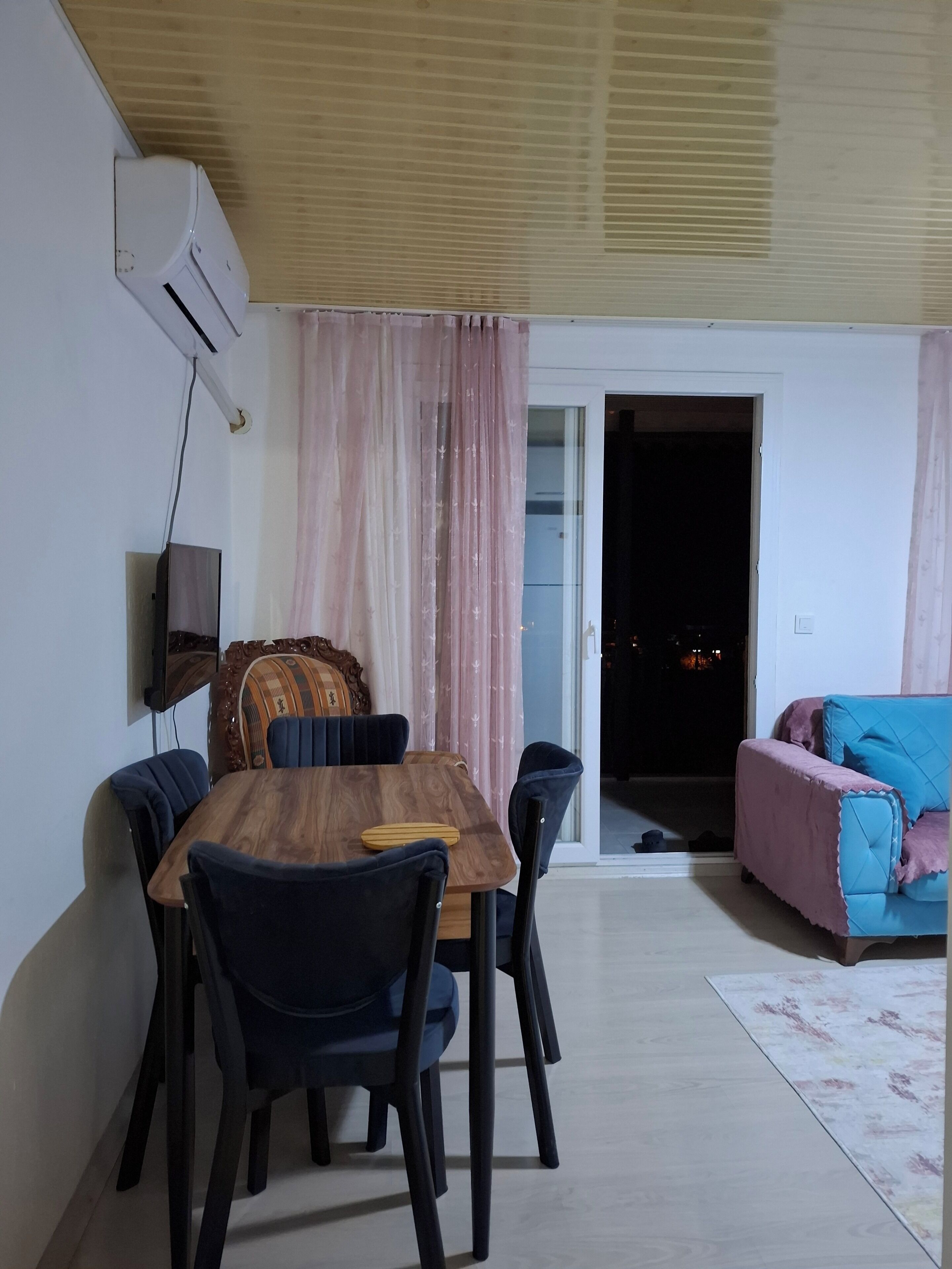 A dining area with a wooden table and chairs, a television mounted on the wall, and a sliding door leading to a balcony.