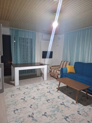 A living room with a white coffee table, a blue sofa with patterned cushions, a TV mounted on the wall, and a patterned rug on the floor. - Demir Apart Otel (Kas)