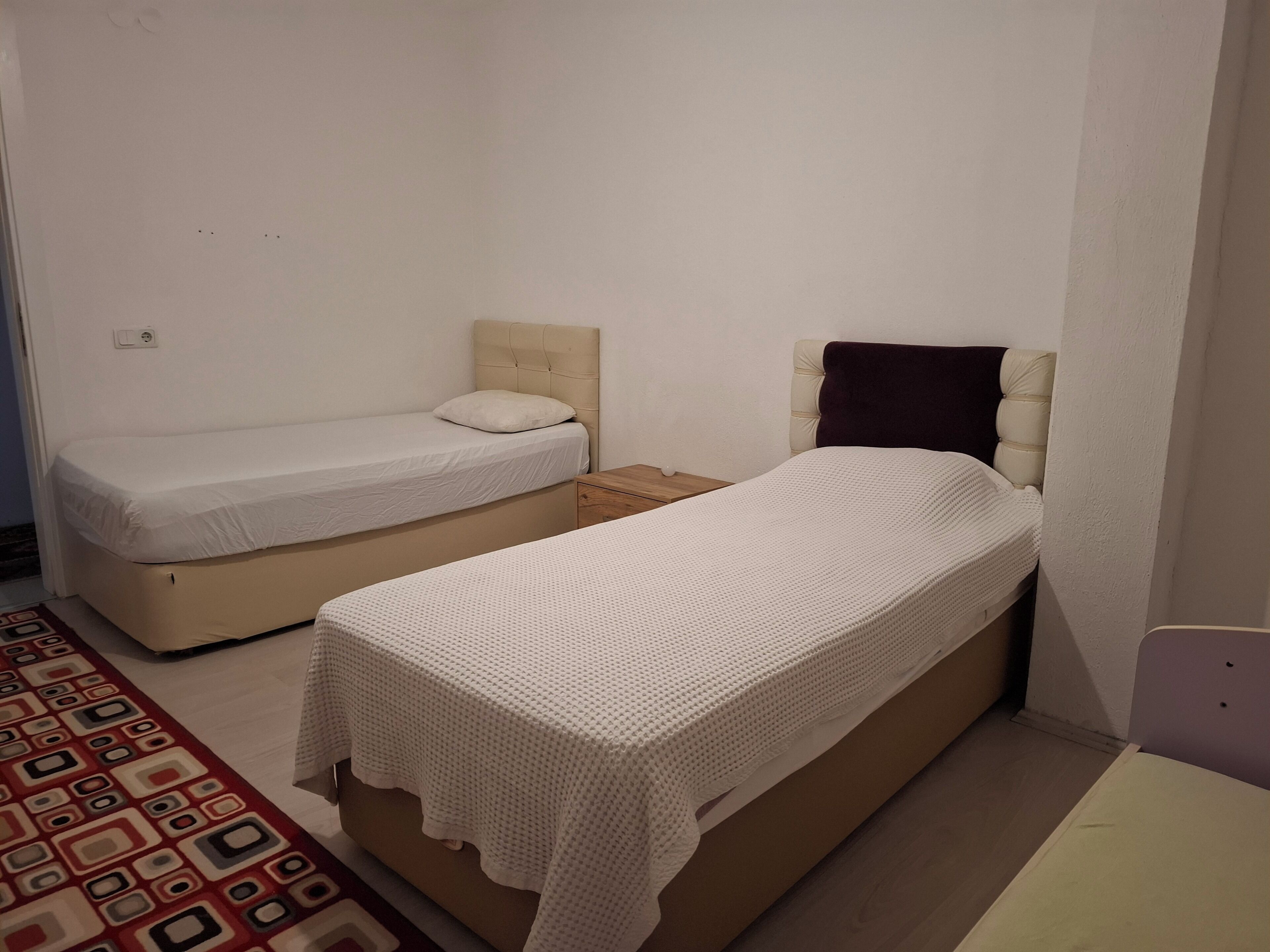 Una habitación pequeña y acogedora con dos camas individuales, una mesita de noche de madera y una alfombra estampada.