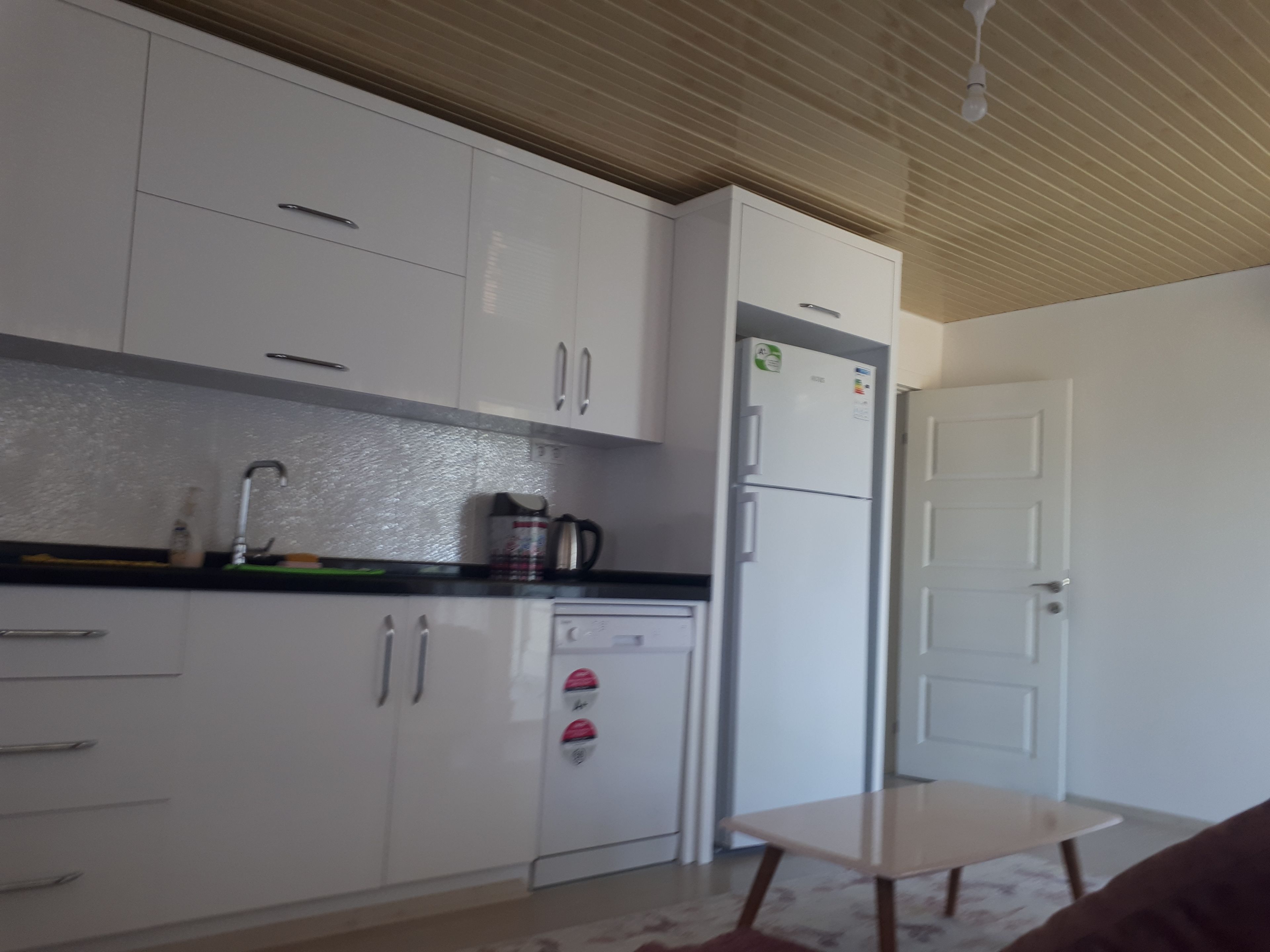Una cocina con armarios blancos, un refrigerador, una cafetera y una pequeña mesa.