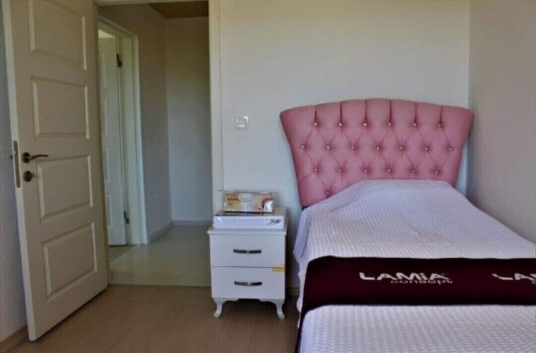 Une chambre à coucher avec une tête de lit rose à motifs capitonnés, une table de chevet blanche avec des tiroirs et un lit recouvert d’une couvre-lit blanche et bordeaux.