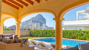 A pool area with lounge chairs, a view of a mountain, and a modern building in the background. - Villa Almendros - PlusHolidays (Calpe)
