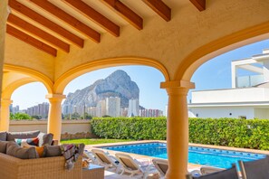A pool area with lounge chairs, a view of a mountain, and a modern building in the background. - Villa Almendros - PlusHolidays (Calpe)