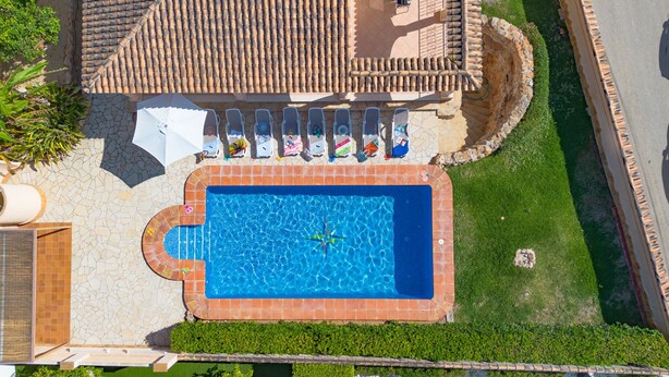 Vista aérea de una piscina con solárium de azulejos, rodeada de un césped cuidado y un muro de piedra.