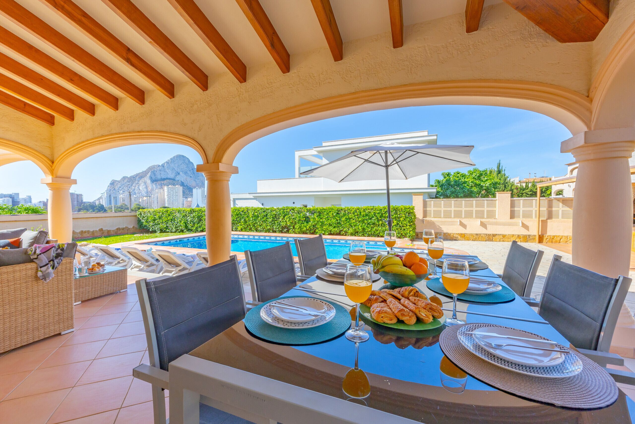 A dining area with a view of a mountain and pool, featuring outdoor seating and a table set for a meal.