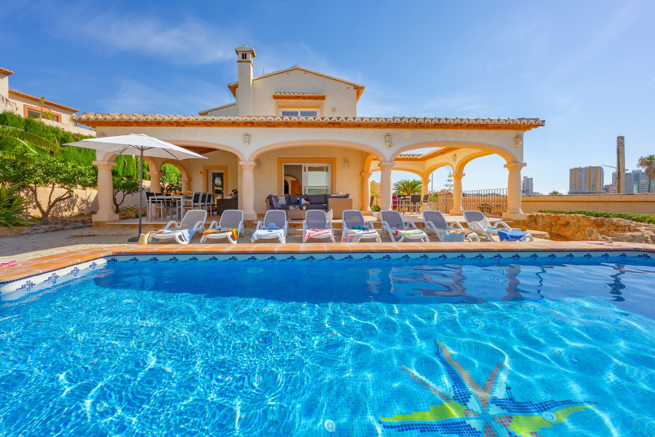 Une piscine avec des chaises longues et des parasols, un patio couvert et un bâtiment doté d’un toit en tuiles.