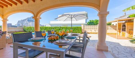 An outdoor dining area with a table set for four, surrounded by white columns, under a wooden ceiling.
