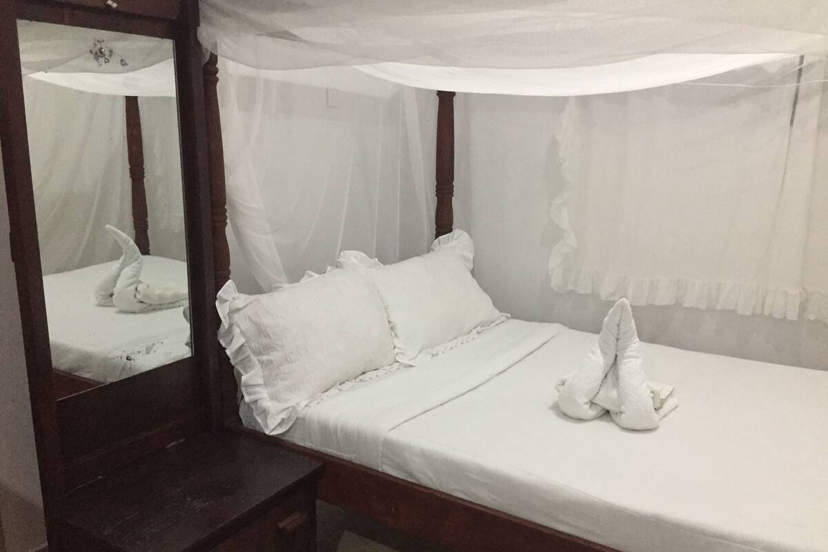 A neatly made bed with white bedding and pillows, a wooden nightstand, and a mirror with a white canopy.