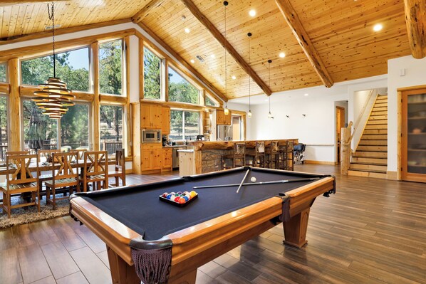 A spacious room with a pool table, wooden furniture, and large windows offering a view of the outdoors.