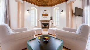 Un salon avec deux canapés blancs, une table basse sur laquelle se trouve un panier de fruits et un verre de vin, une cheminée et un téléviseur fixé au mur.