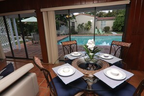 Spiseområde med et glasbord og stole, udsigt til poolen og en terrasse med udendørs møbler.
