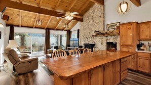 A spacious living area with a large wooden dining table, a fireplace, and a stone wall. There are ceiling fans, a sofa, and a TV in the room. - Scenic Retreat #1772 (Big Bear City)