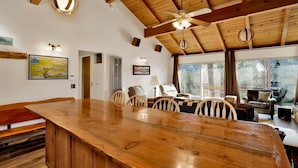 A spacious room with a wooden dining table, chairs, a sofa, a ceiling fan, and large windows offering a view of trees. - Scenic Retreat #1772 (Big Bear City)