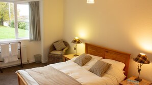 Een net opgemaakt bed met een houten hoofdbord, twee bedlampen, een rieten fauteuil met kussens, een radiator en een raam met gordijnen.