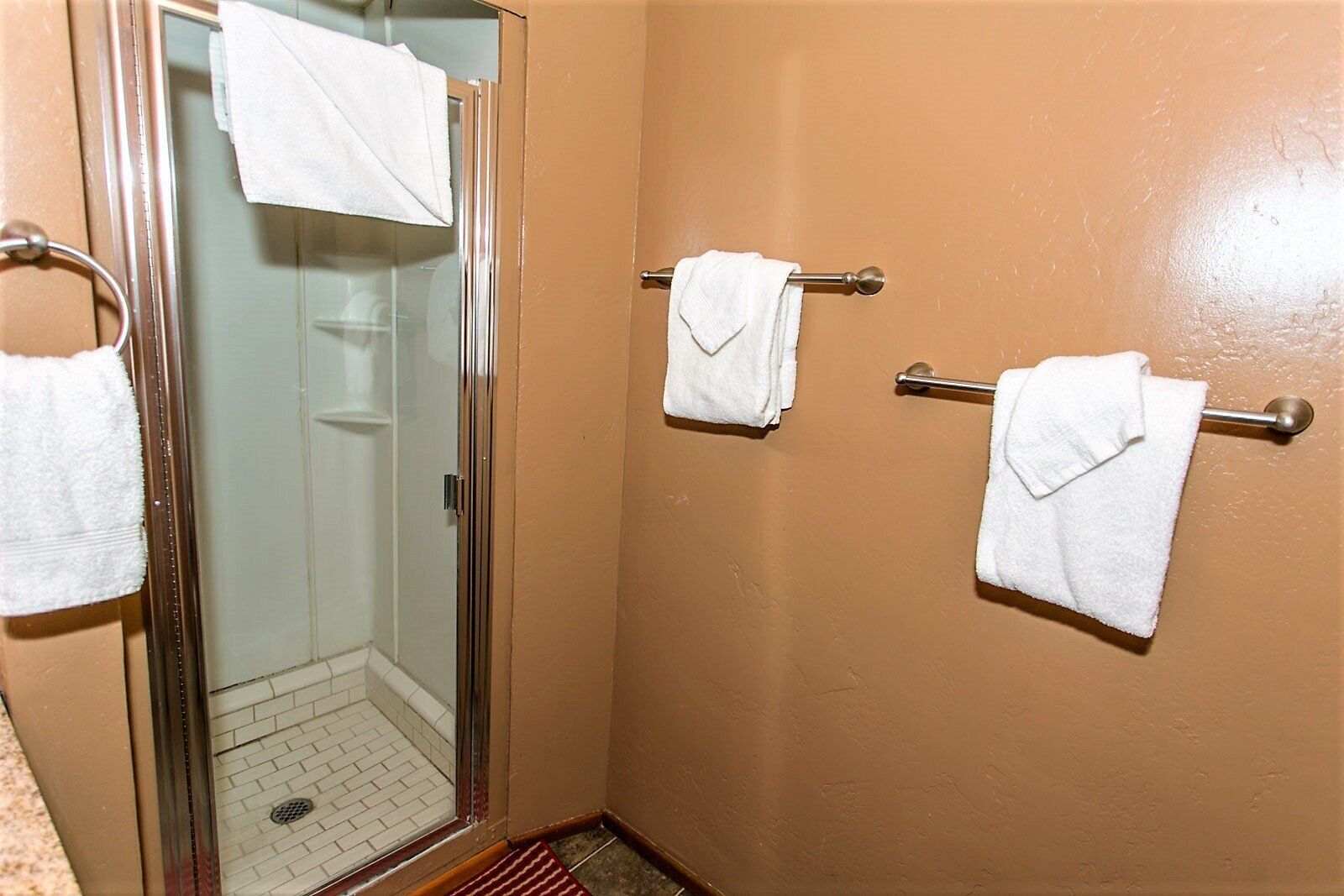 A bathroom with a glass shower door, two towel bars with white towels, and a red doormat.