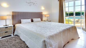 A spacious bedroom with a large bed, two bedside lamps, a decorative wall piece, and a view of a swimming pool through a large window. - Villa Licinia - Three Bedroom Villa, Sleeps 6 (Guia)