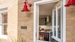 Fachada de ladrillo con ventana y puerta, un accesorio de luz rojo, y un vistazo a una cocina y zona de estar en el interior.