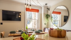 Comedor con mesa de madera dispuesta para cuatro personas, un espejo grande y una ventana con cortinas naranjas.