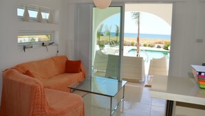 A modern living room with a glass table, a sofa, and a view of a beach and pool. - Imagine Your Family Renting a Luxury Holiday Villa Close to Sotira’ Main Attractions (Sotira)