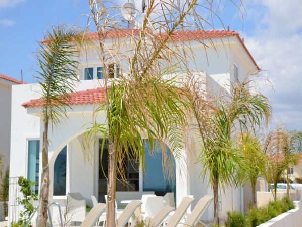 A modern building with a white facade and red-tiled roof, featuring a pool area with lounge chairs and palm trees.