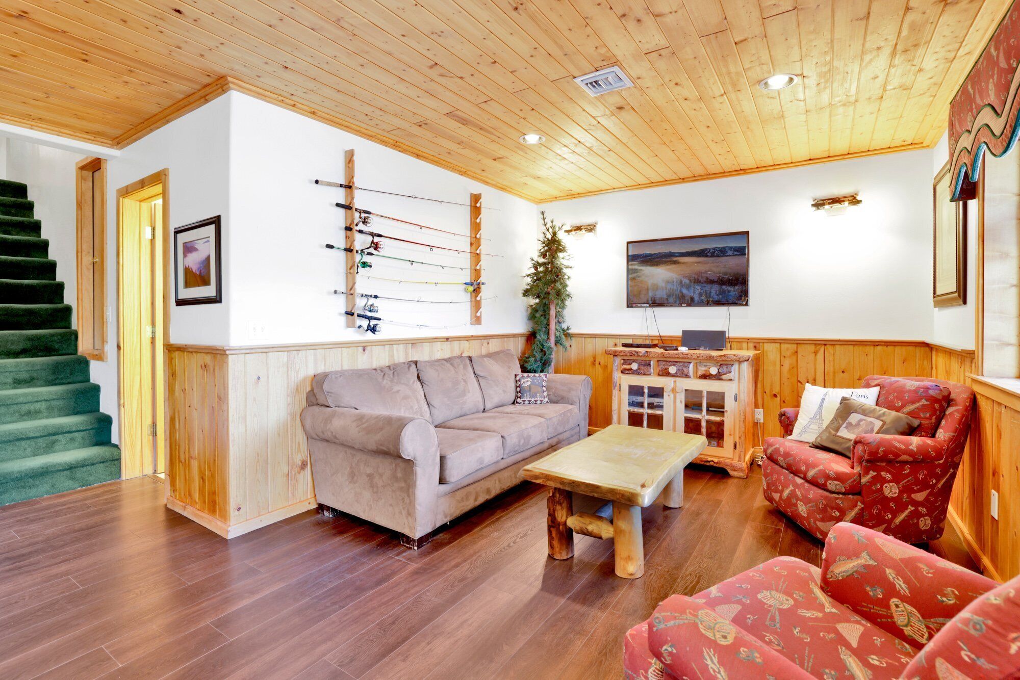 Una sala de estar acogedora con techo de madera, un sofá, un sillón rojo con estampado, una mesa de centro de madera y una colección de cañas de pescar colgadas en la pared.