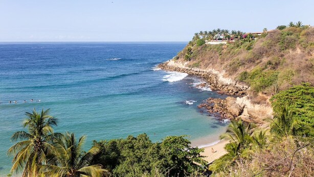Une vue côtière comprenant une plage, une côte rocheuse et un bâtiment doté d’un jardin, situé sur le versant d’une colline.