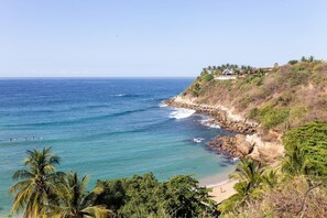 A coastal view with a beach, rocky shoreline, and a building with a garden on a hillside.