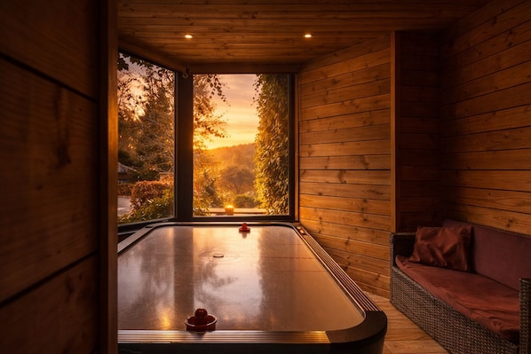 A sauna room with a wooden interior, a glass door leading to a view of trees and a sunset, and a wicker sofa.