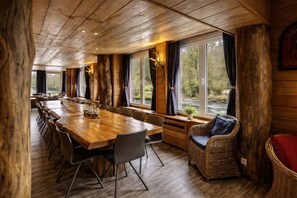 A wooden interior with a long dining table, wicker furniture, and large windows with curtains.
