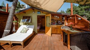 A wooden cabin with a balcony, a small kitchen area, and a sleeping area with a bed and two chairs.